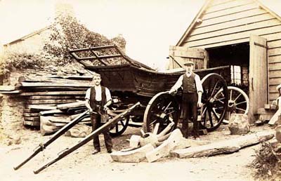 The photograph shows the cartwrights workshop with two men, dressed in white shirts, waistcoats and cord trousers, standing in front of a large wooden wagon with big wheels. One man is holding a paint pot and brush in his hands.