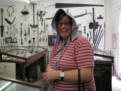 A smiling woman tries on a lavender coloured old-fashioned cotton bonnet in Clun Museum. In the backgroun are the Museum's displays of farming implements.  