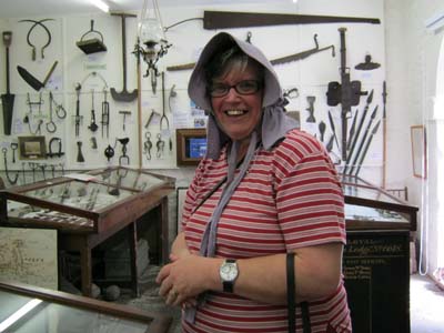 A smiling woman tries on a lavender coloured old-fashioned cotton bonnet in Clun Museum. In the backgroun are the Museum's displays of farming implements. A smiling woman tries on a lavender coloured old-fashioned cotton bonnet in Clun Museum. In the backgroun are the Museum's displays of farming implements.