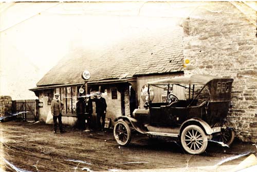 A vintage car, with a folding roof, is standing in front of the garage. It is high off the ground. To the left are the petrol pumps.