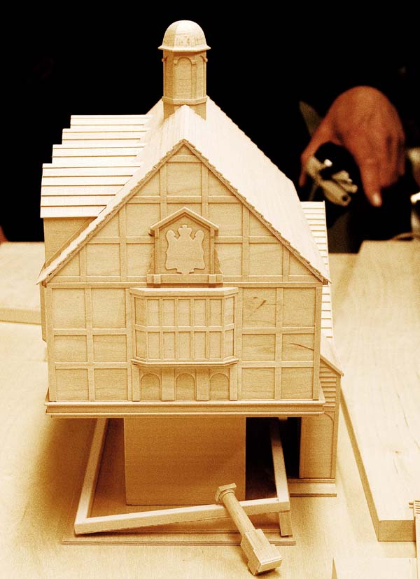 A pale coloured wooden model of an old house with pitched roof and gable windows stands on a table.