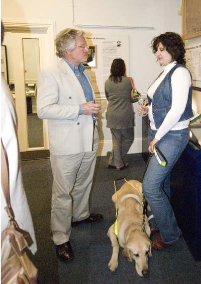Maggie, wearing blue jeans and white jersey, is talking to Paul Rew who is wearing a light coloured suit. Maggie's golden labrador guide dog is lying on the floor between them.