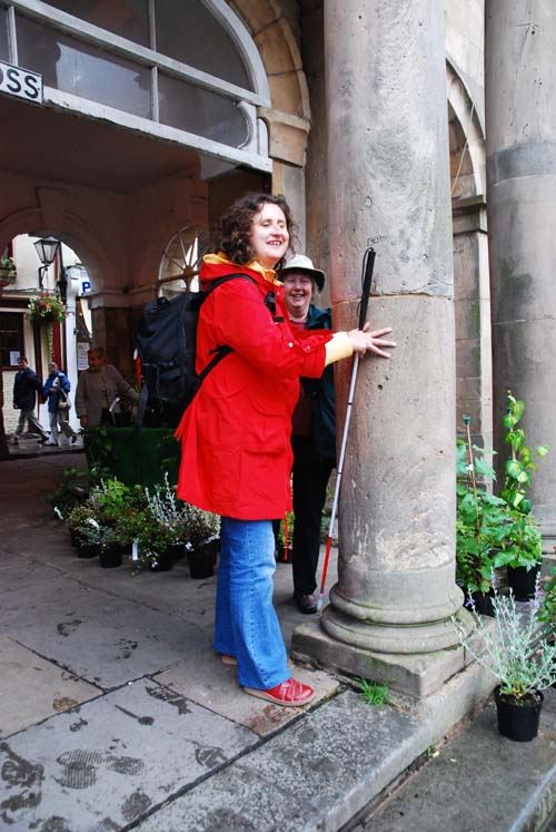 A young woman in a red coat explores the cream stone column of the 18th century Buttercross. In front of her there are two steps and behind her some plants for sale on a market stall.