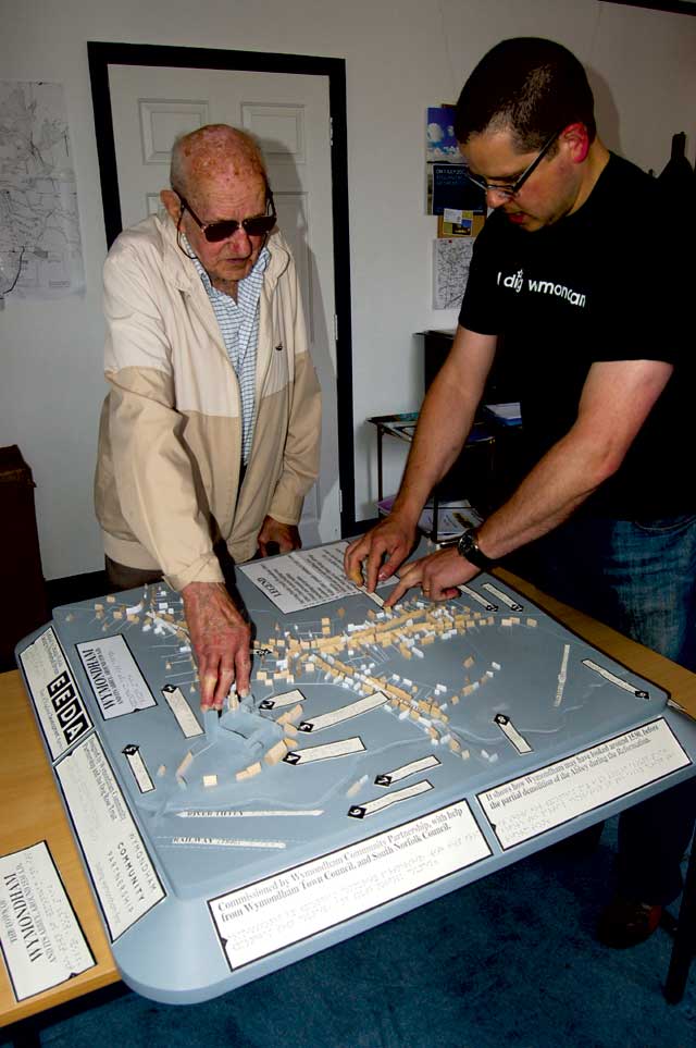 An elderly blind man in a fawn jacket and dark glasses is feeling the buildings on the model with his right hand. To  his left is a younger man in a black t-shirt running his hand over the Braille which has been created temporarily with swell-paper.